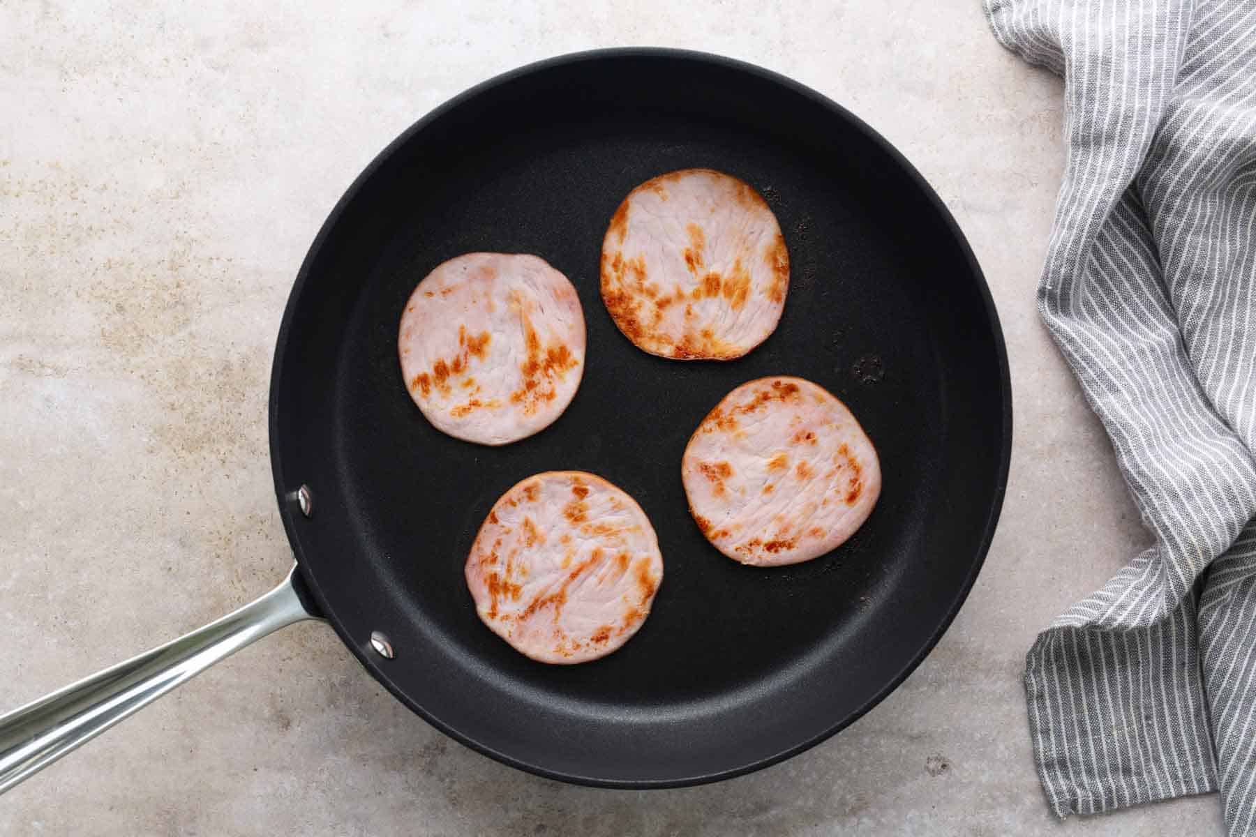 Four round slices of Canadian bacon are cooking in a black skillet, positioned on a light countertop next to a gray striped cloth—perfectly prepared and ready to be layered onto a classic eggs benedict.