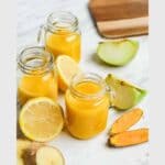 Three small glass jars of yellow juice are surrounded by lemon slices, ginger, turmeric, and green apple pieces on a marble surface. Text reads: "ginger turmeric shot," "zesty," and "refreshing.