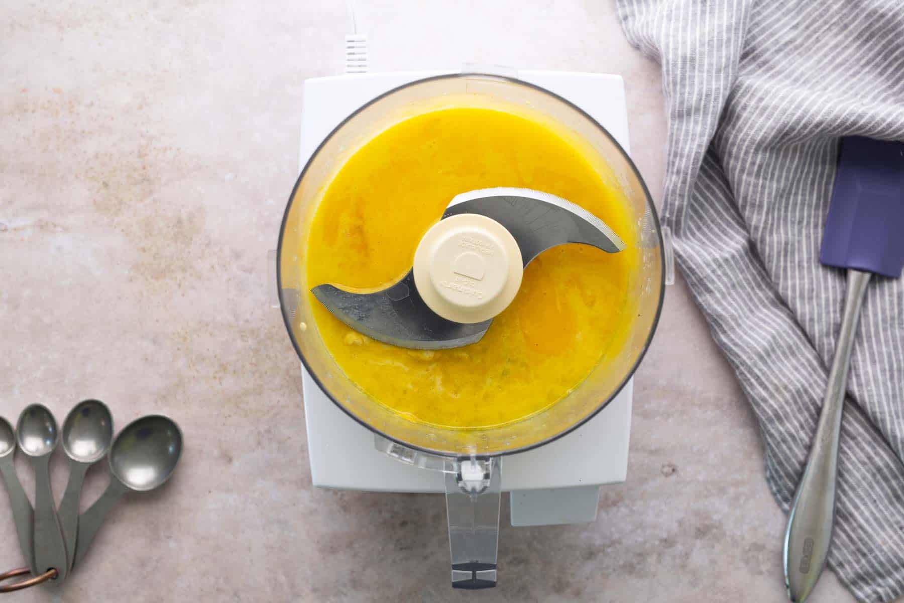 Overhead view of a food processor with a yellow liquid mixture inside, surrounded by measuring spoons, a striped towel, and a spatula on a light countertop.