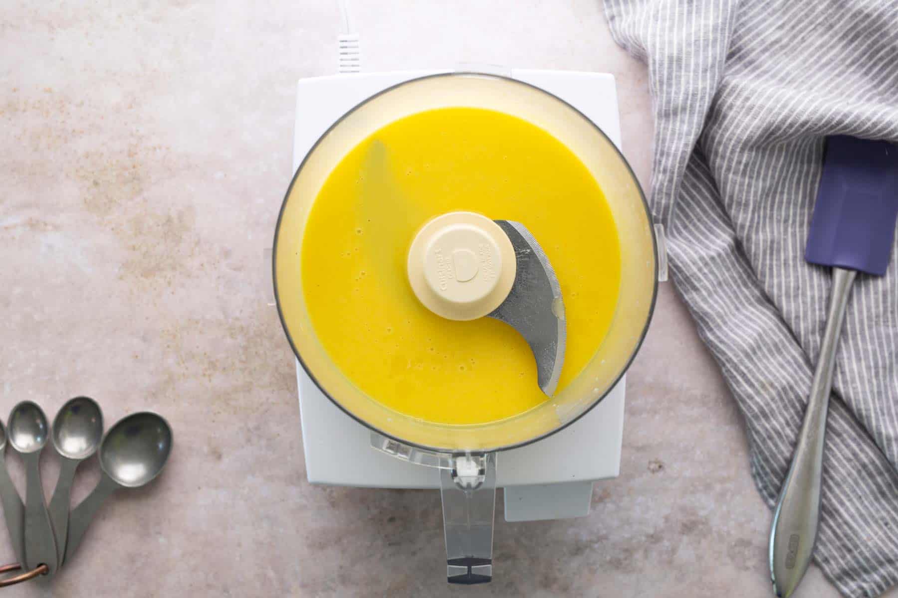 Overhead view of a food processor with a yellow liquid mixture inside, sitting on a light countertop next to measuring spoons, a striped towel, and a purple spatula—perfect for preparing silky hollandaise sauce for classic eggs benedict.