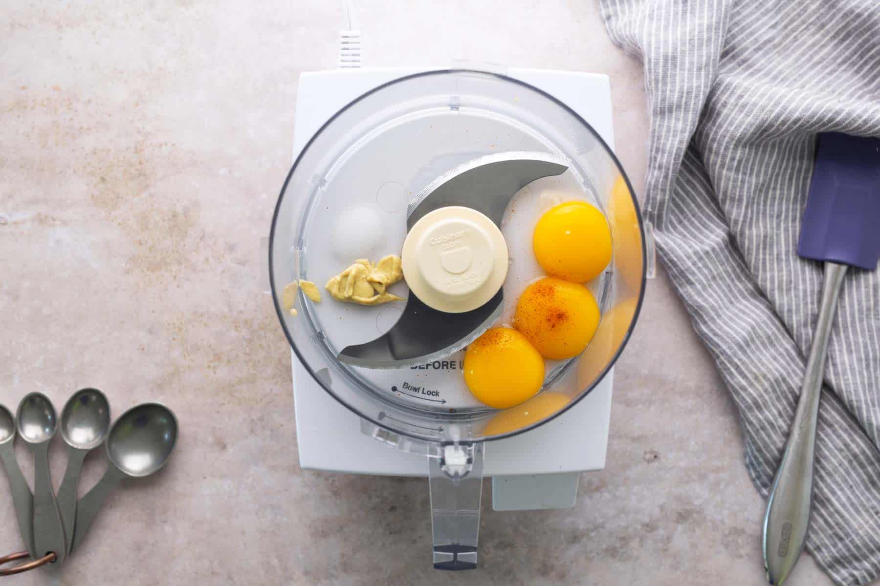 Top view of a food processor bowl with three egg yolks, mustard, and seasoning inside, on a countertop next to a striped towel, measuring spoons, and a spatula.