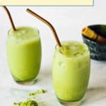 Two glasses of iced matcha latte with metal straws sit on a white surface. Matcha powder and a small black bowl with a bamboo whisk are nearby.