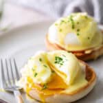 Two eggs Benedict on English muffins with hollandaise sauce and chopped chives, one egg cut open and yolk running, served on a plate with a fork.