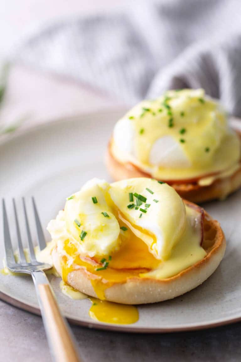 Two eggs Benedict on English muffins with hollandaise sauce and chopped chives, one egg cut open and yolk running, served on a plate with a fork.
