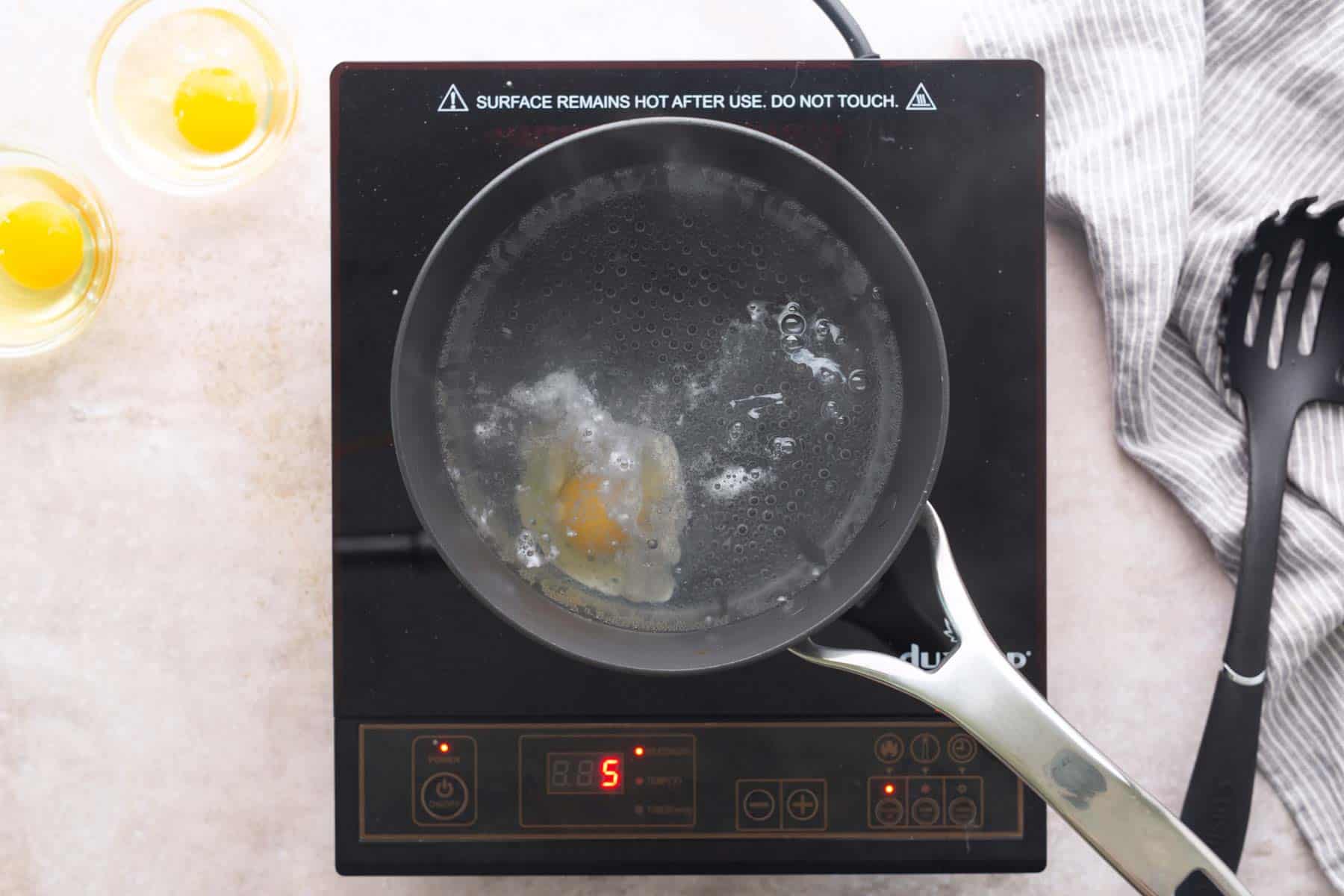 A saucepan with a poaching egg cooks on an induction cooktop set to level 5; two cracked eggs are in bowls nearby, ready to be transformed into classic eggs benedict.