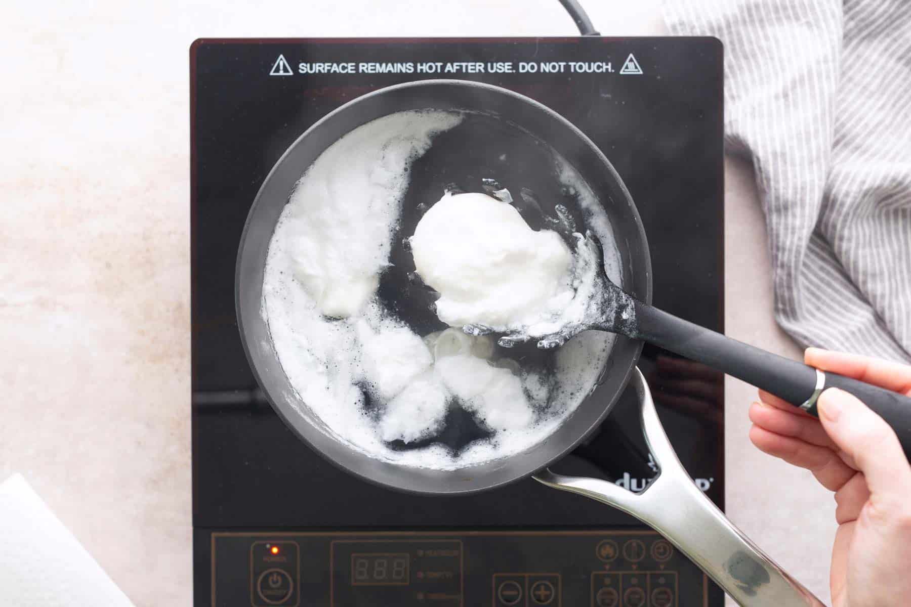 A person uses a slotted spoon to skim white foam from boiling water in a saucepan on an electric cooktop.