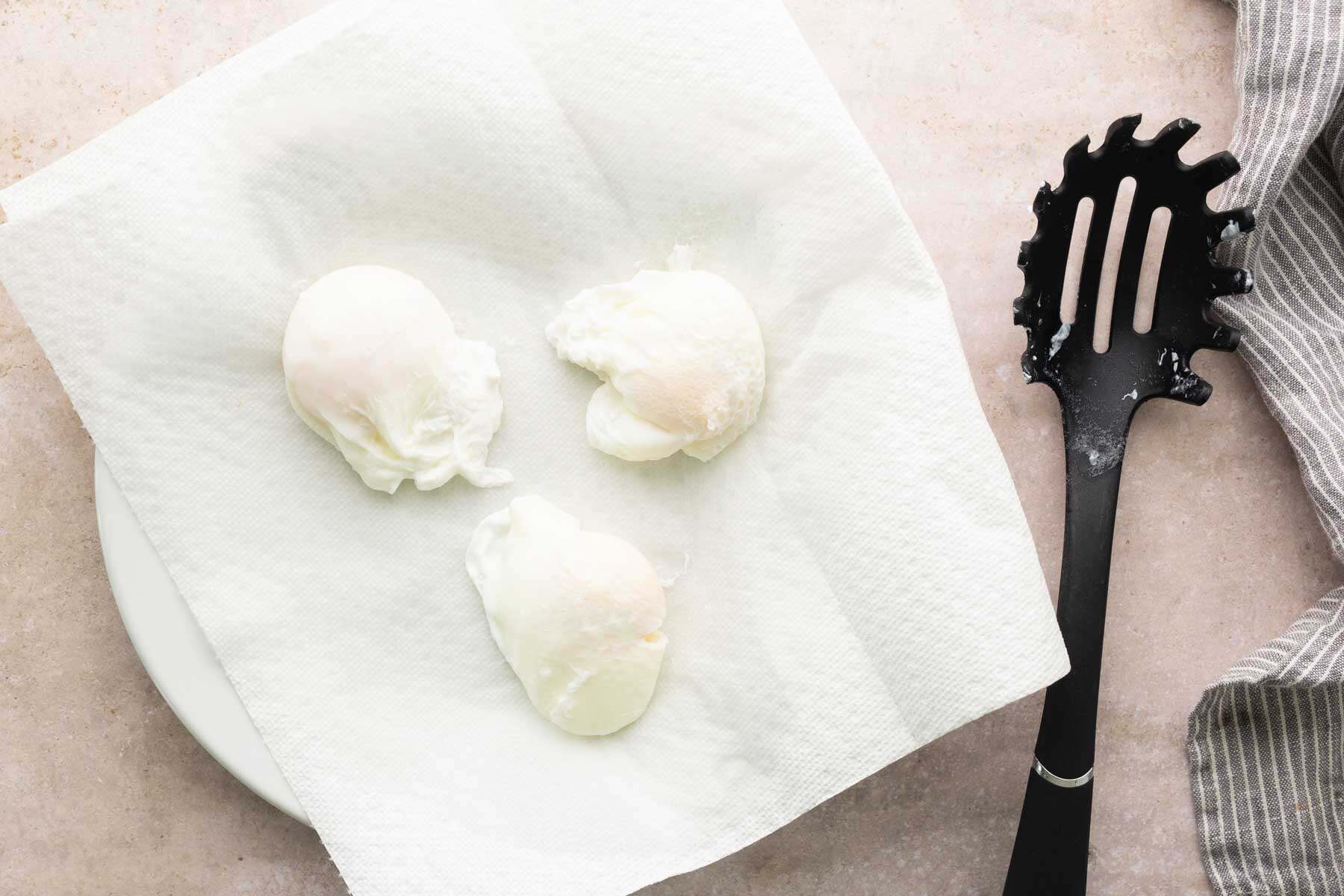 Three poached eggs rest on a paper towel-lined plate next to a black slotted spoon and a gray striped cloth on a light countertop.