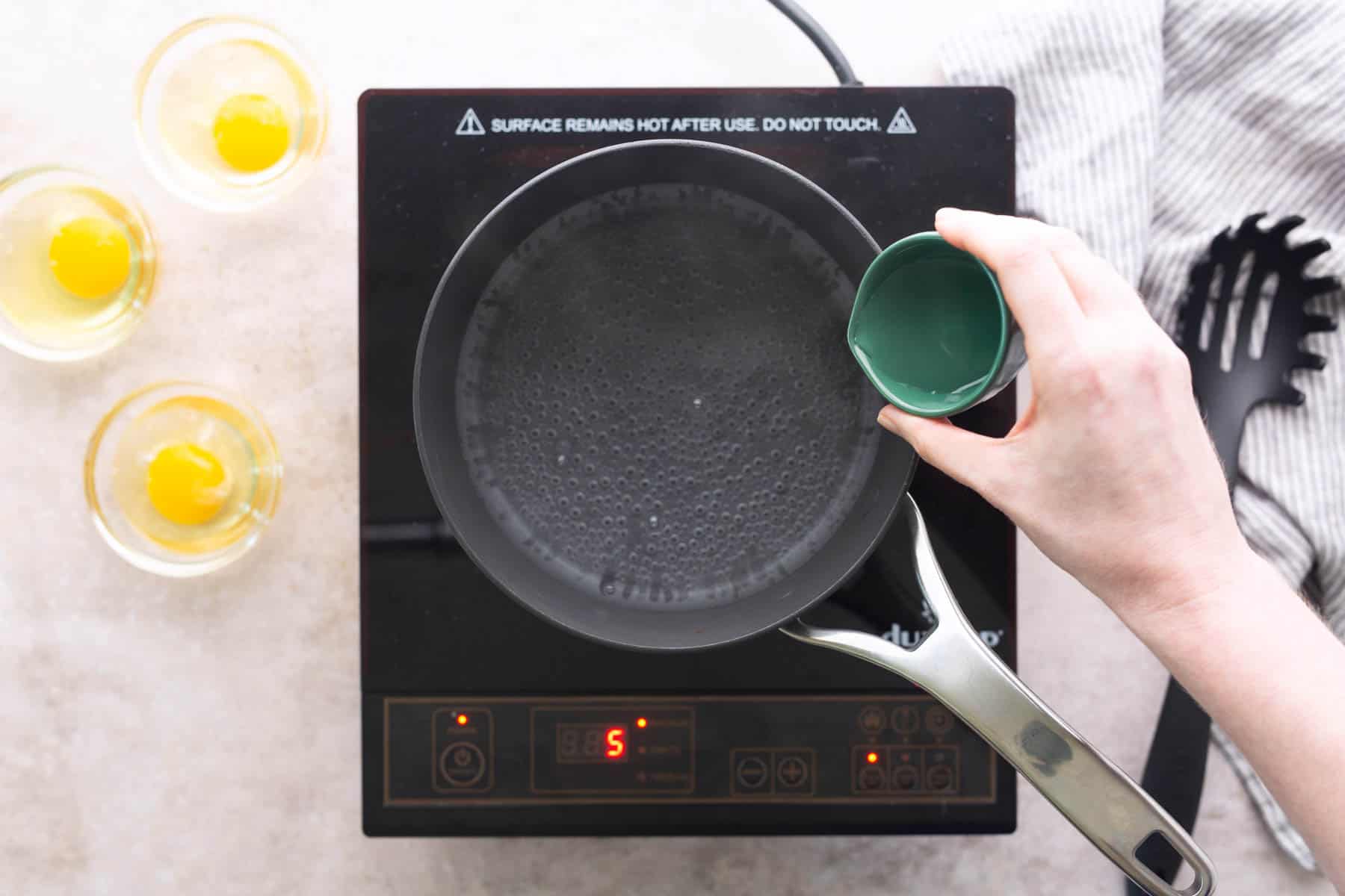 A hand pours liquid from a small green cup into a pot of boiling water on an electric stovetop, with three cracked eggs in cups nearby.
