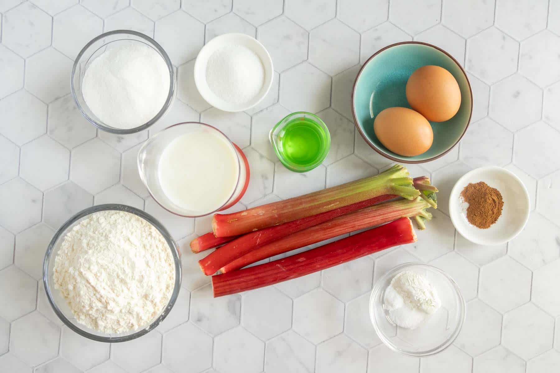 Various baking ingredients, perfect for making rhubarb muffins, including flour, sugar, eggs, milk, rhubarb stalks, and spices are arranged on a white hexagonal tile countertop.