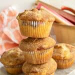 Four cinnamon sugar muffins are stacked on a marble surface, with a basket of rhubarb and a pink patterned cloth in the background—an inviting scene perfect for showcasing your favorite rhubarb muffins.