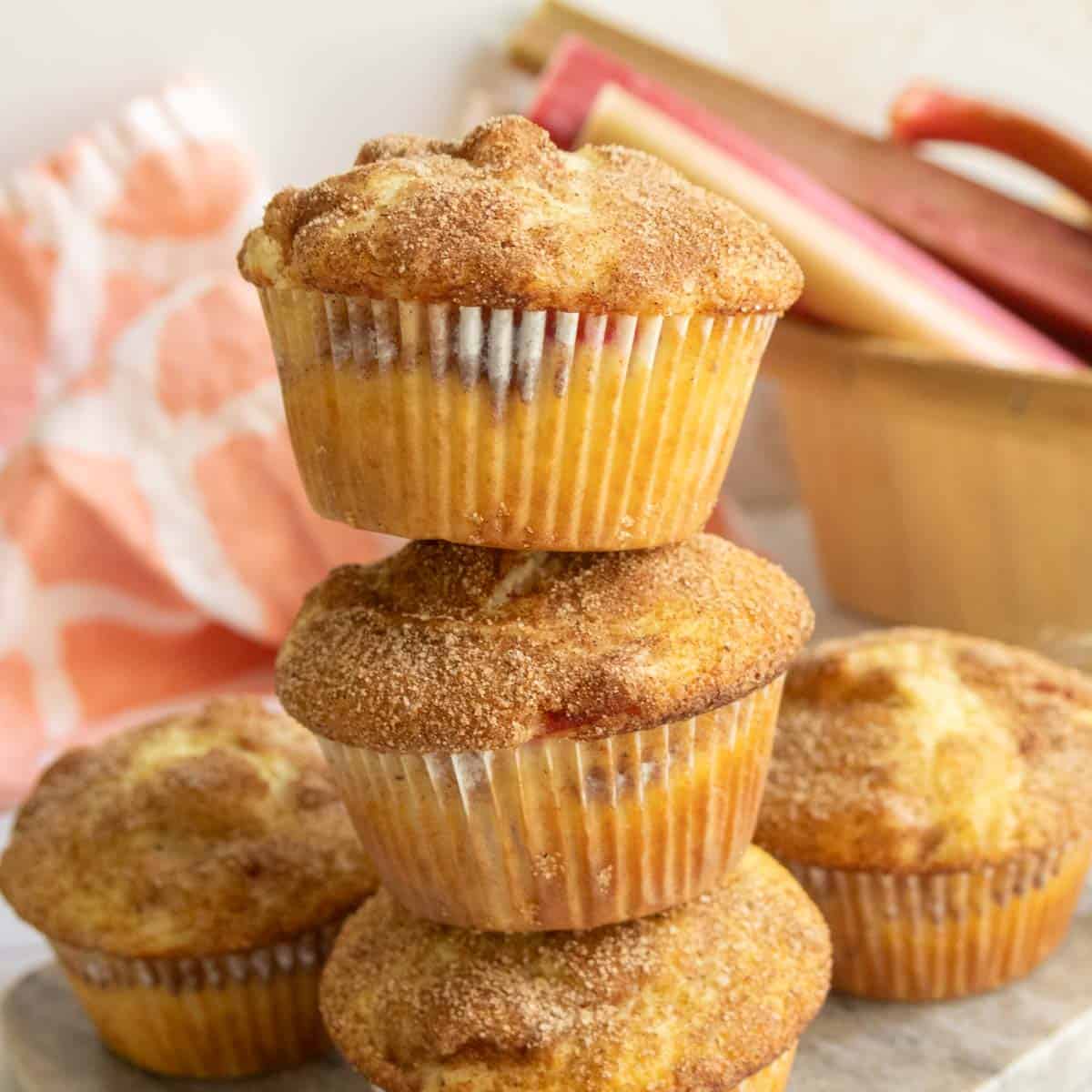 A stack of four cinnamon sugar muffins in paper liners, perfect for breakfast recipes, with a basket of rhubarb and a pink kitchen towel in the background.