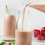 A glass bottle is being filled with strawberry almond milk, with strawberries and mint leaves on the table, and another filled bottle in the background.