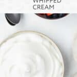 A bowl of vegan whipped cream sits next to a bowl of mixed berries and a spoon on a white surface.
