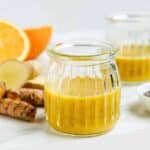 A small glass jar filled with yellow chia seed wellness shot, with ginger, orange, and turmeric pieces in the background.