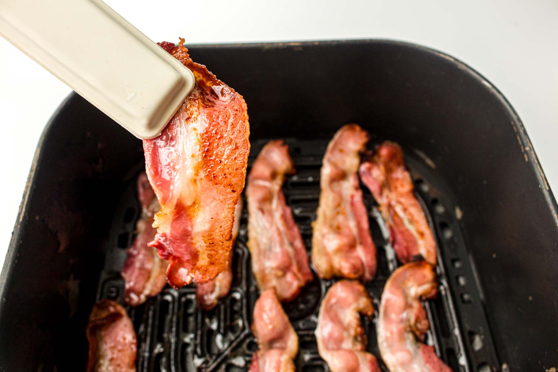Tongs holding up a slice of cooked bacon over an air fryer basket.