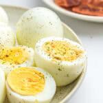 A plate of air fryer hard boiled eggs, some halved to show the yolks, seasoned with black pepper and herbs. Blurred in the background is a plate of sliced toast or bread.