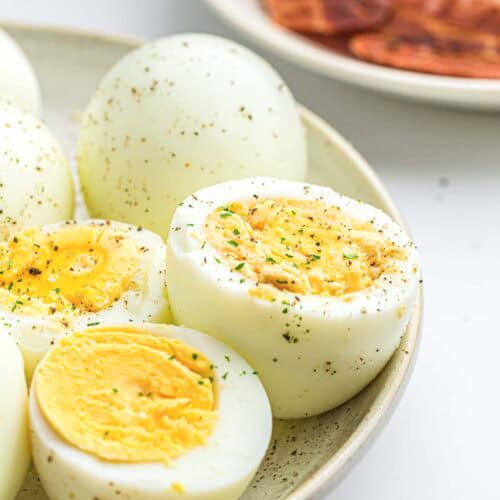 A plate of air fryer hard boiled eggs, some halved to show the yolks, seasoned with black pepper and herbs. Blurred in the background is a plate of sliced toast or bread.