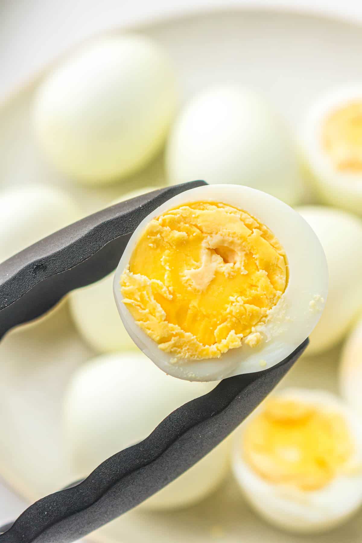 Black tongs hold a halved air fryer hard boiled egg with a bright yellow yolk; several whole eggs are blurred in the background on a plate.