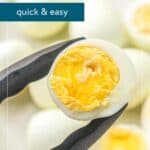 A pair of tongs holds a halved hard-boiled egg with a yellow yolk; several whole eggs are blurred in the background.