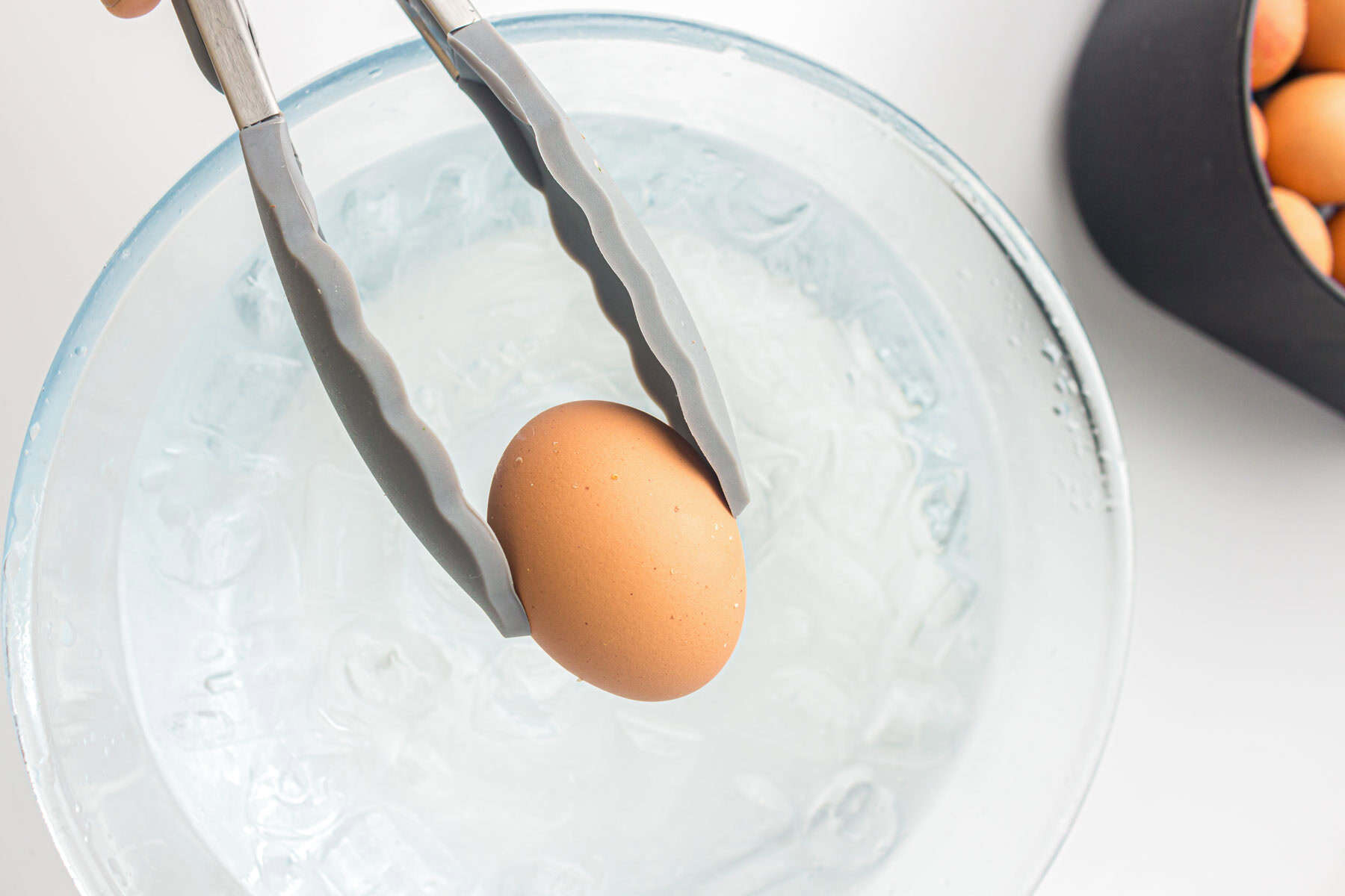 A pair of tongs holds a brown egg above a glass bowl filled with ice water, demonstrating the process of making air fryer hard boiled eggs; more eggs are visible in a container nearby.