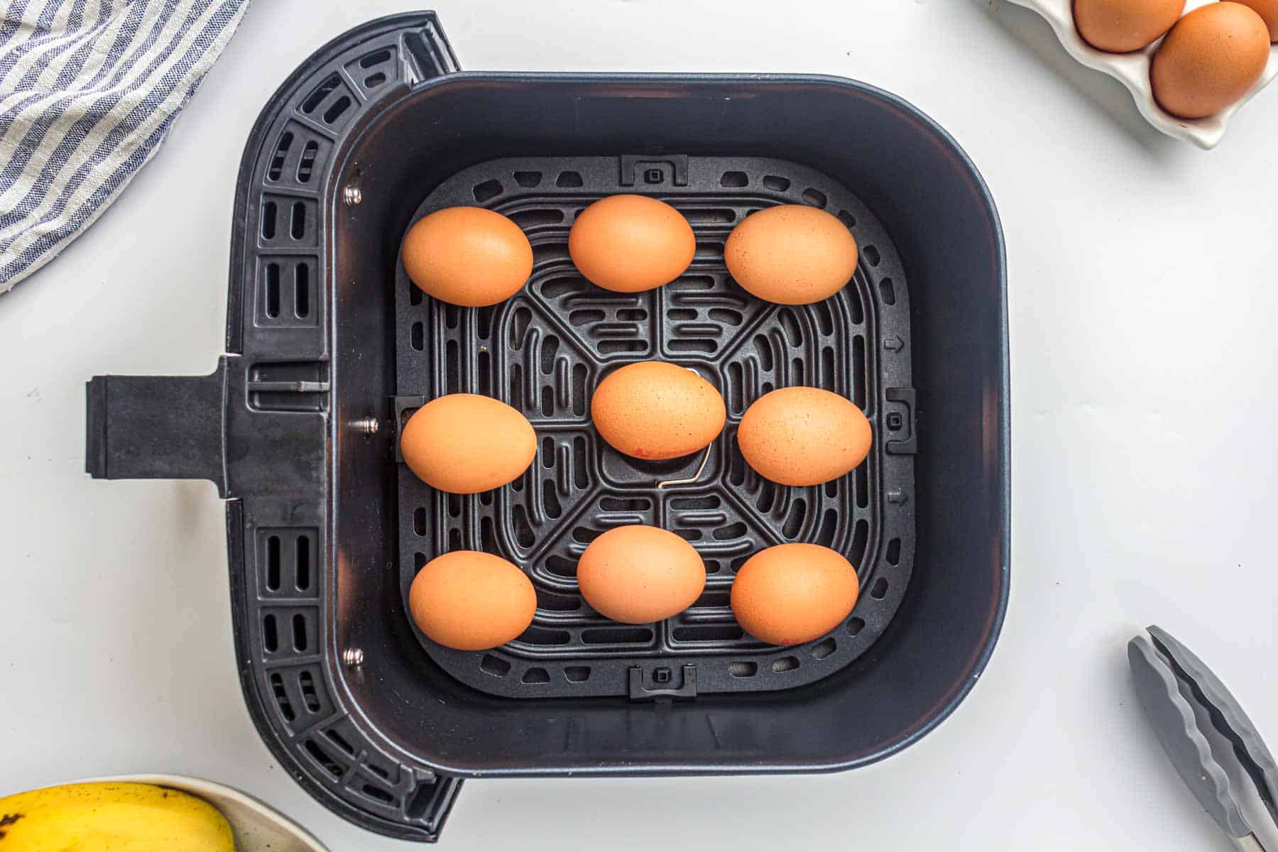 Nine brown eggs are arranged inside the basket of a black air fryer, ready to make easy air fryer hard boiled eggs, with kitchen items like tongs and a striped towel visible around it.