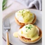 Two classic eggs benedict topped with silky hollandaise sauce and fresh chives rest on toasted English muffins, served on a plate with a fork beside them.