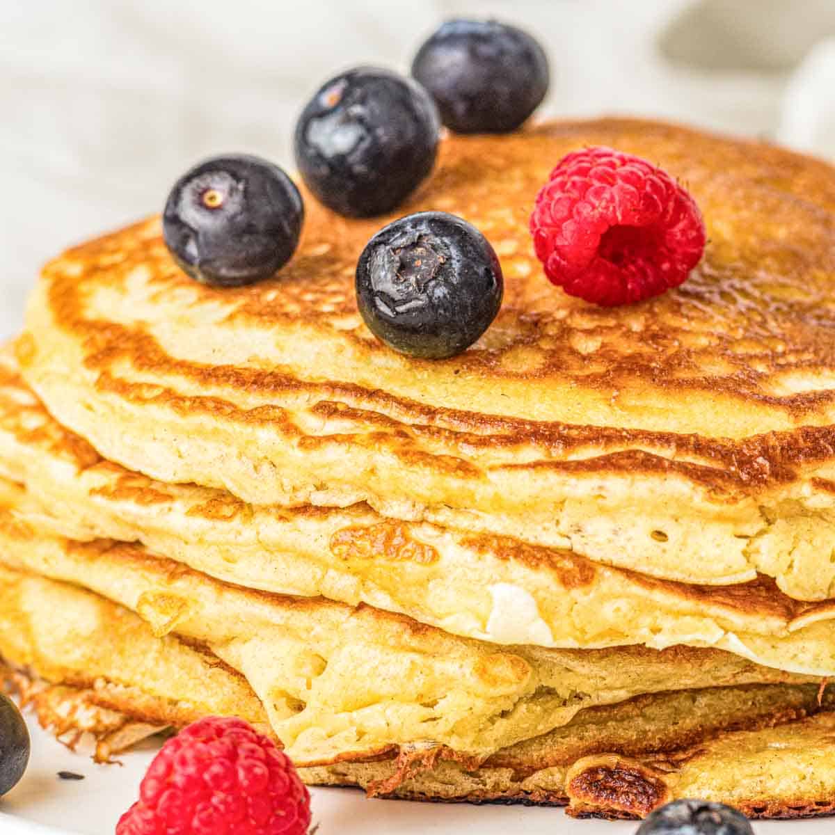 A stack of pancakes topped with fresh blueberries and a raspberry, with more berries scattered nearby.