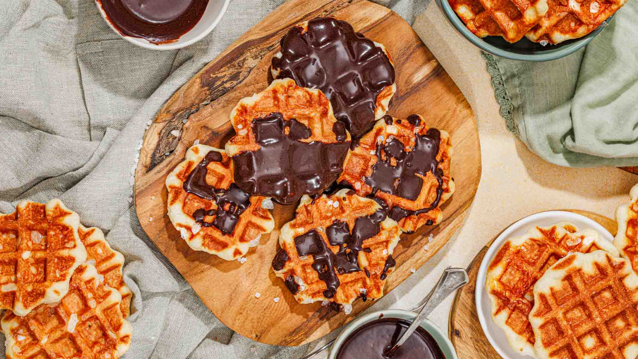 A wooden board with waffles, some partially dipped in chocolate, surrounded by bowls of chocolate sauce and more waffles on a table with a green cloth.