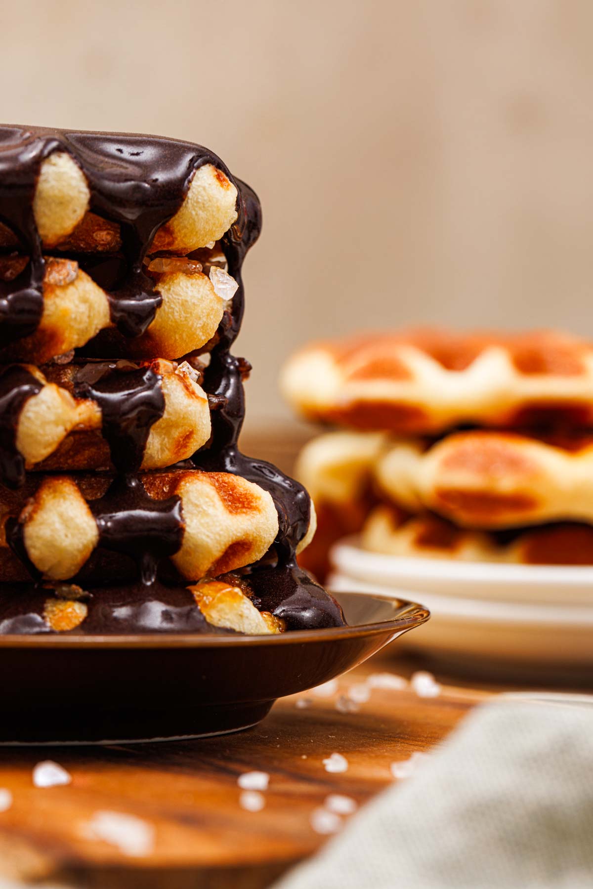 Stack of waffles drizzled with chocolate sauce on a dark plate, with another stack of plain waffles on a white plate in the background.