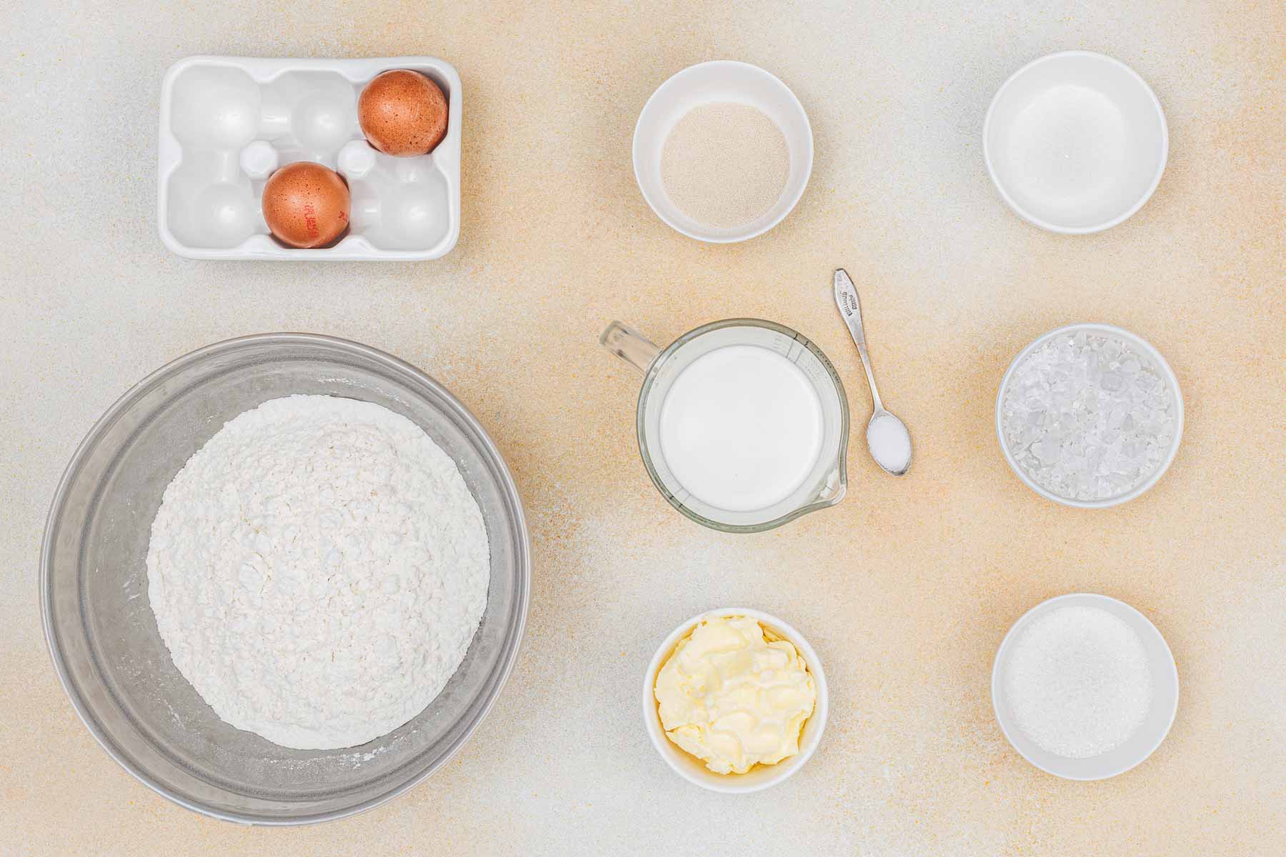 Top view of baking ingredients including eggs, flour, milk, yeast, sugar, salt, and butter, each placed in separate bowls on a light-colored surface.