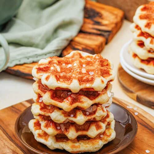 A stack of four golden brown waffles topped with coarse sugar sits on a dark plate on a wooden board, with more waffles and breakfast items in the background.