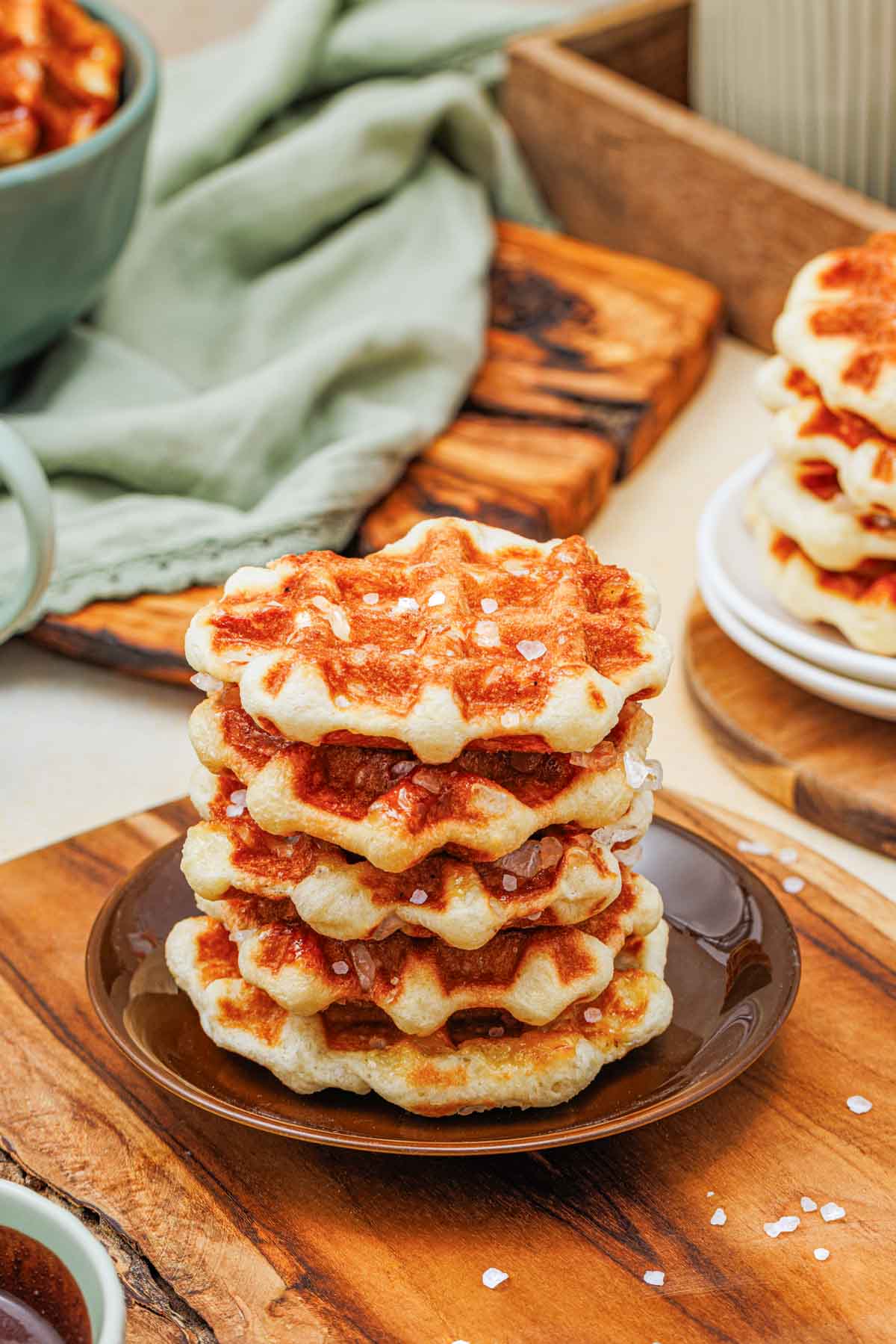 A stack of four golden brown waffles topped with coarse sugar sits on a dark plate on a wooden board, with more waffles and breakfast items in the background.