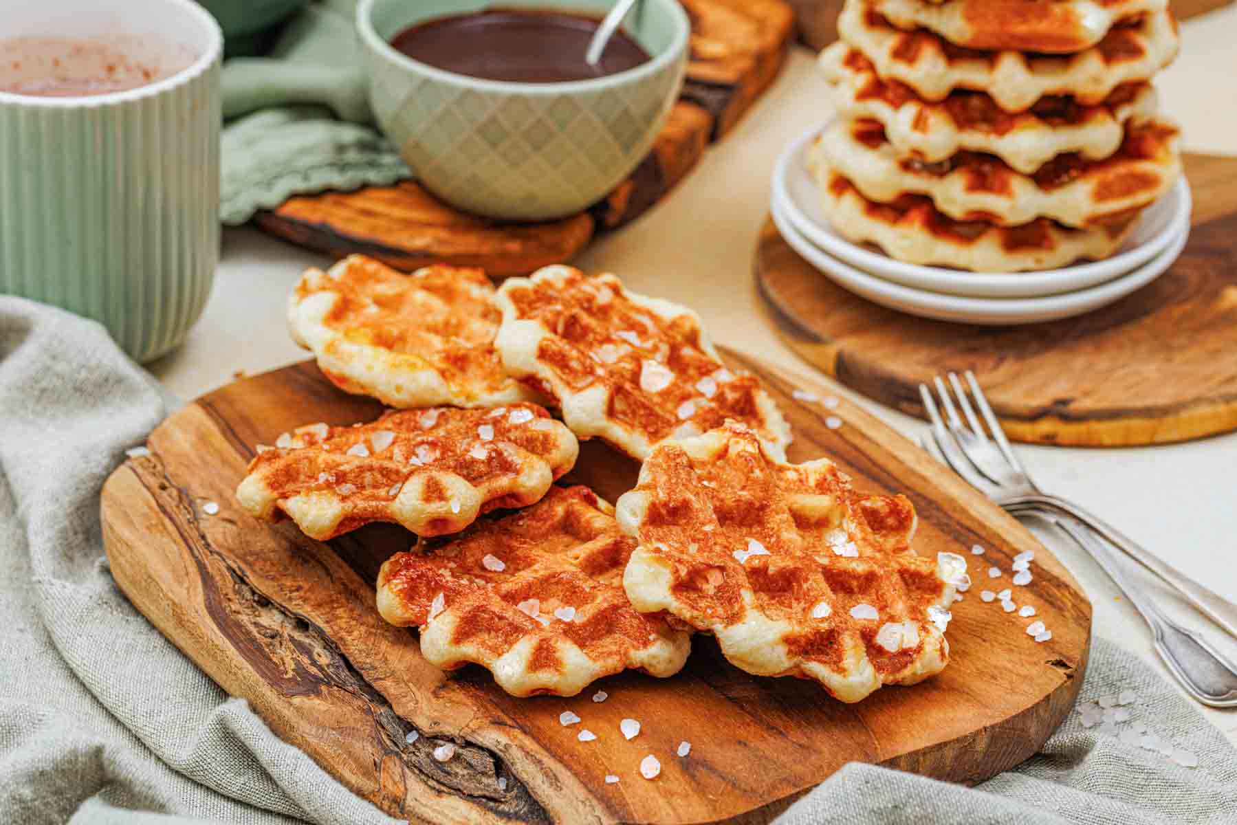 Several golden waffles with coarse sugar sit on a wooden board, with stacked waffles, forks, mugs, and bowls of chocolate sauce in the background.