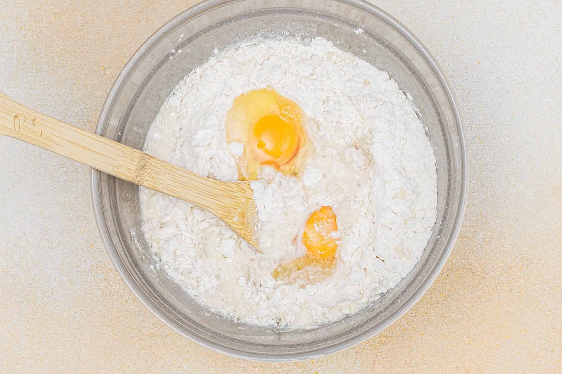 A clear bowl with flour, two cracked eggs, and a wooden spoon, ready for mixing ingredients on a light surface.