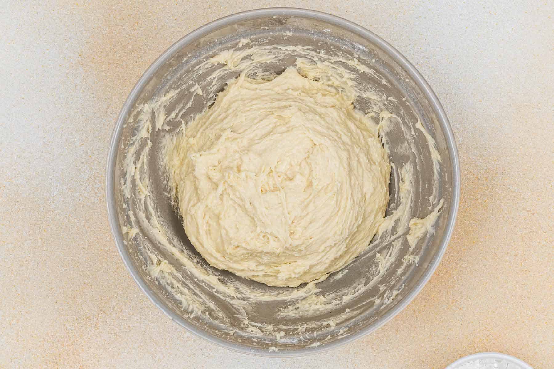 A glass bowl containing thick, creamy dough sits on a light-colored surface.