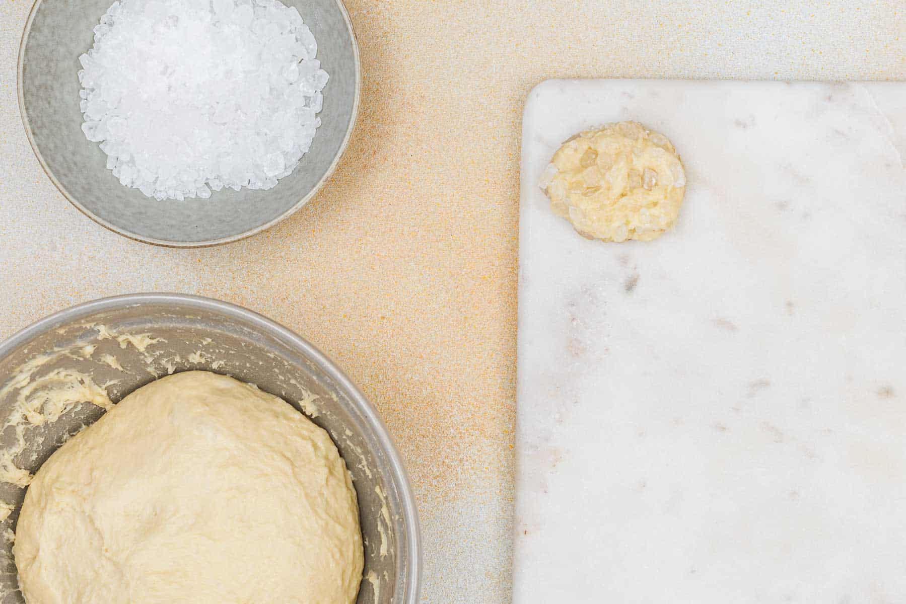 A bowl of coarse salt, a bowl of bread dough, and a small dough piece sit on a white marble surface.