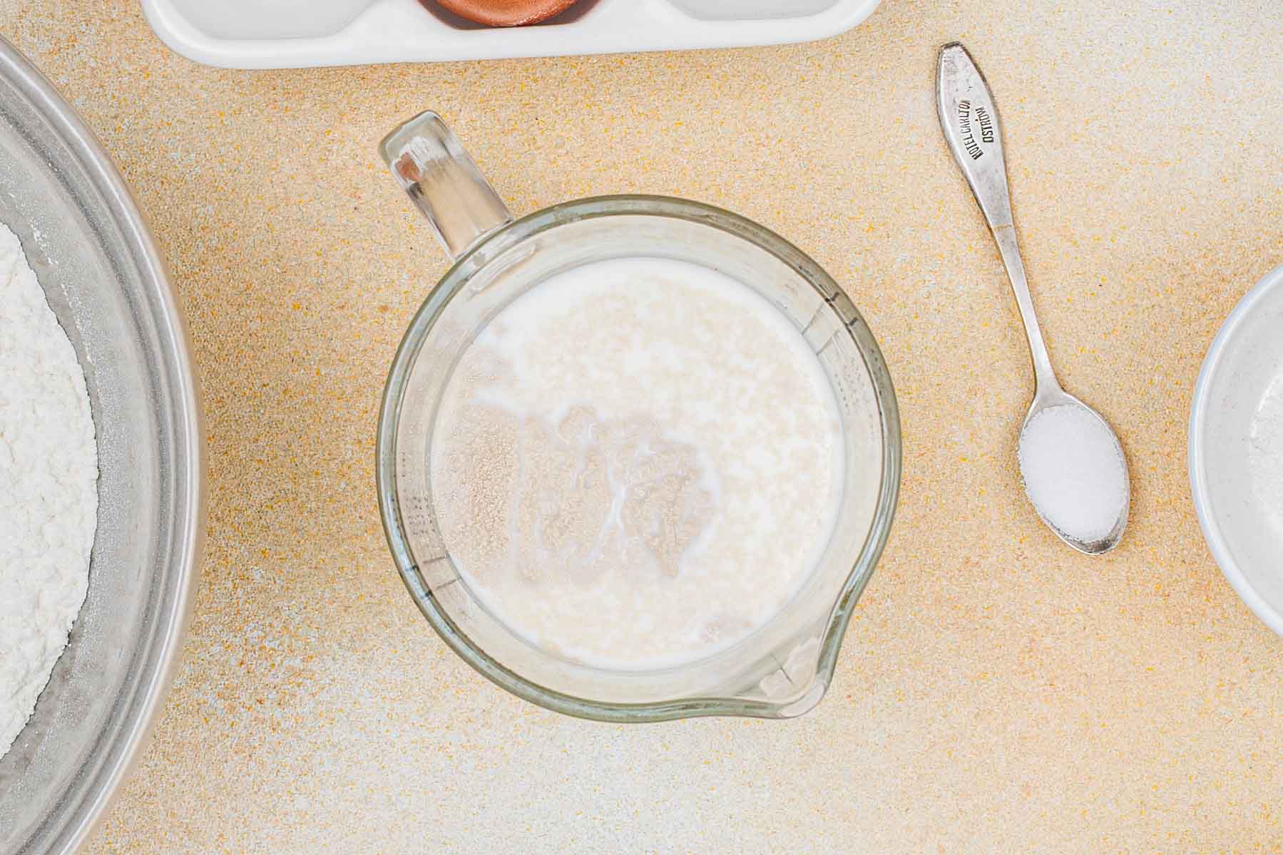 A glass measuring cup with milk and yeast mixture sits on a countertop, surrounded by bowls of flour and a spoonful of salt.