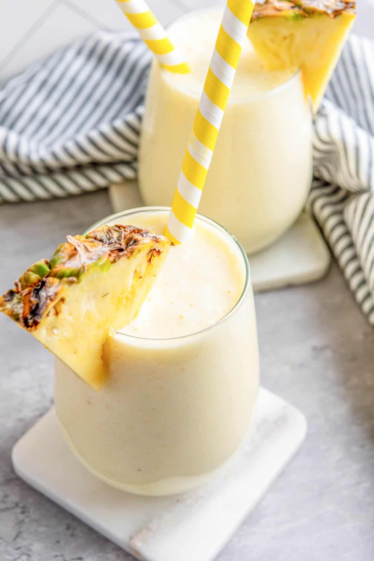 Creamy pineapple smoothies in two glasses with yellow striped straws.
