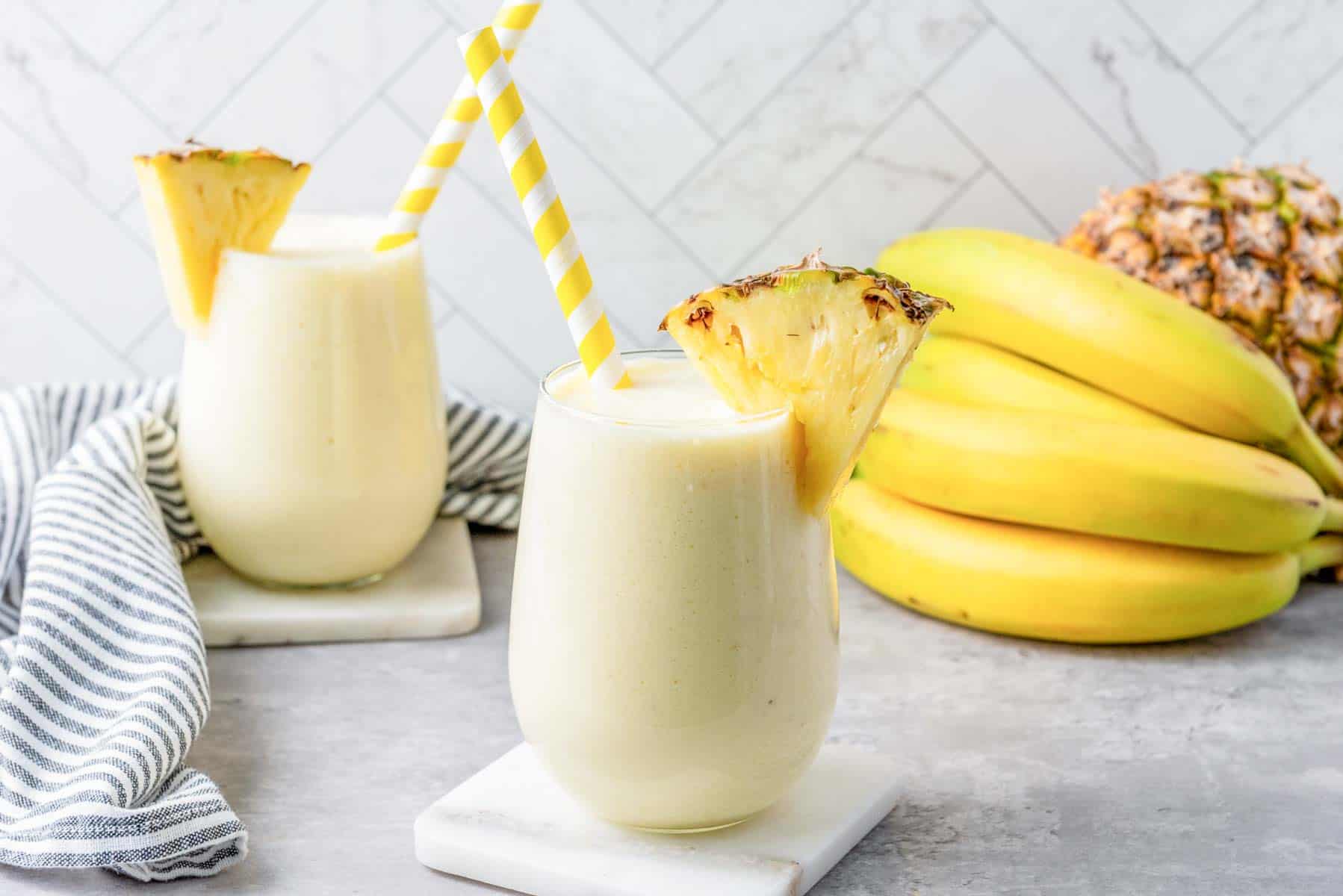 Pineapple smoothie in two glasses, with straws and pineapple slices sticking out of them.