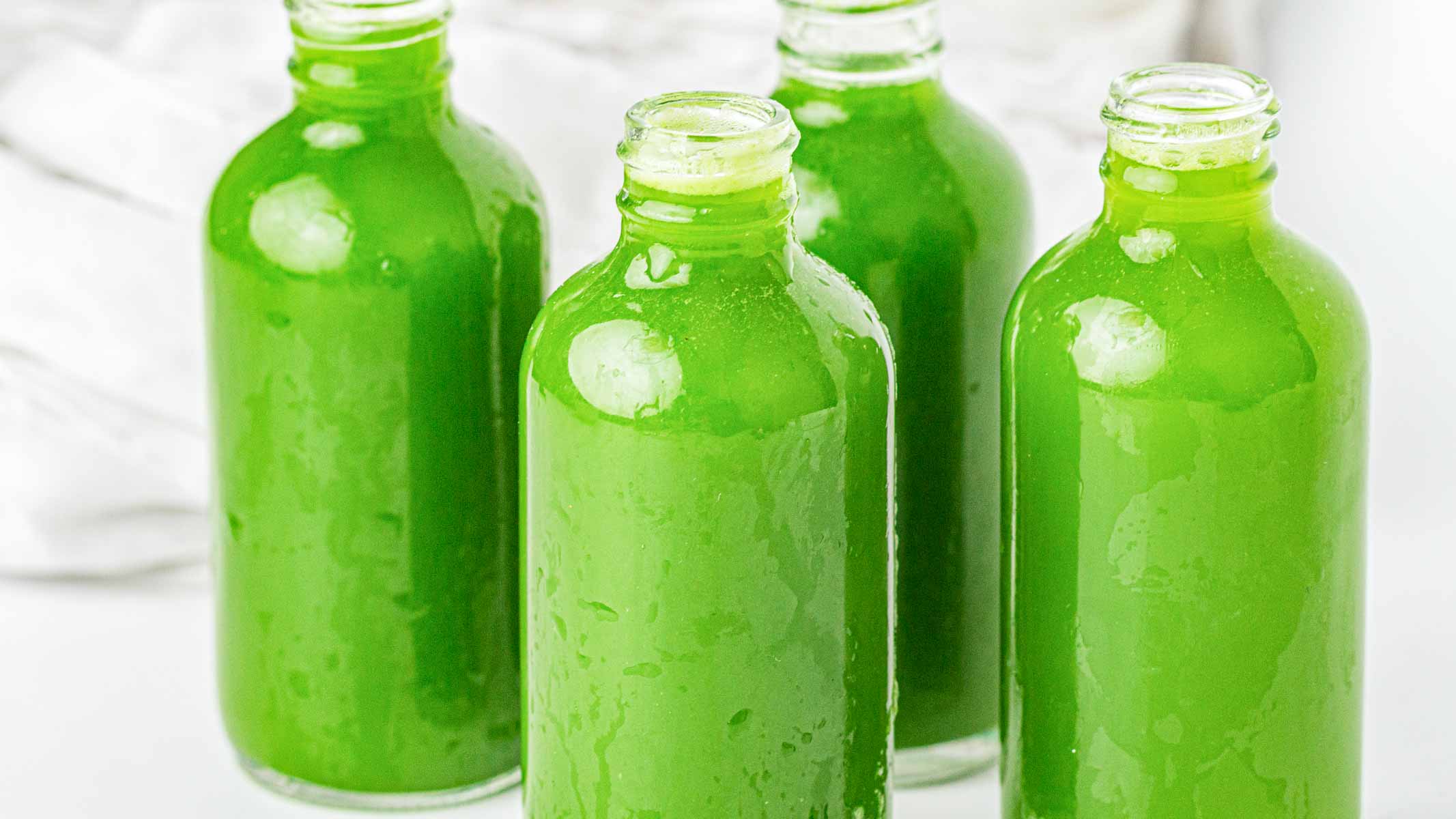 Four clear glass bottles filled with a bright green liquid are arranged closely together on a white surface.