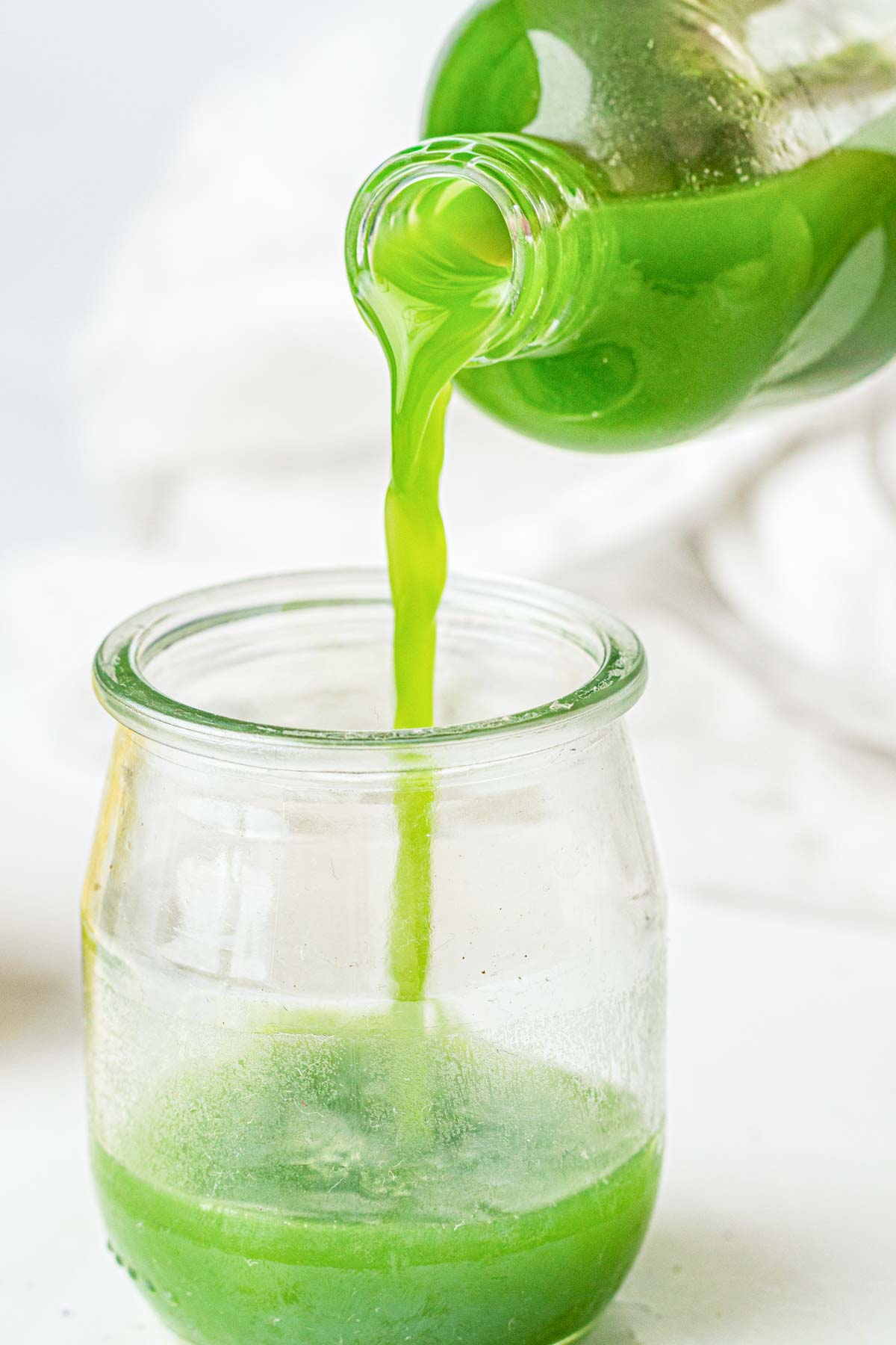Green juice is being poured from a bottle into a small, clear glass jar on a white surface with a blurred background.