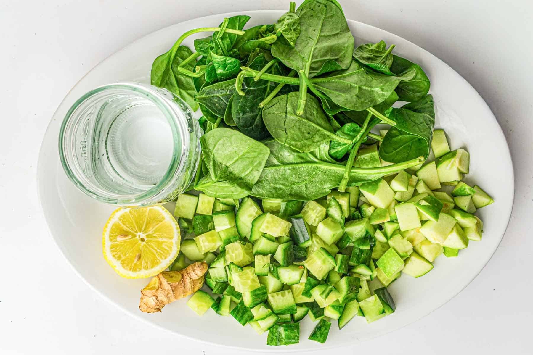 A white plate with spinach leaves, diced cucumber, diced green apple, a lemon half, a piece of ginger, and a glass of water.