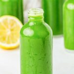 A clear glass bottle filled with bright green juice sits on a white surface, with more bottles and half a lemon visible in the background.