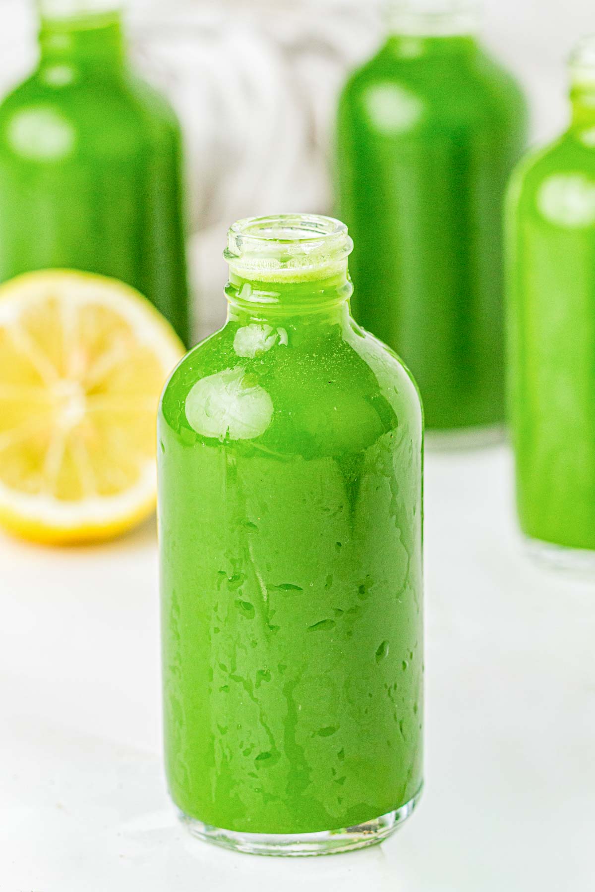 A clear glass bottle filled with bright green juice sits on a white surface, with more bottles and half a lemon visible in the background.
