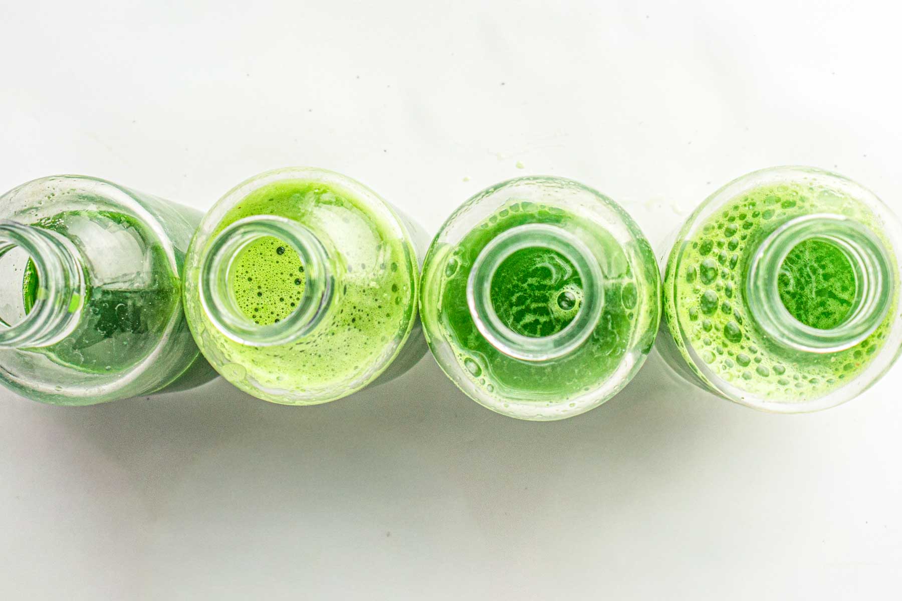 Top view of four glass bottles filled with green juice, arranged in a row on a white background.
