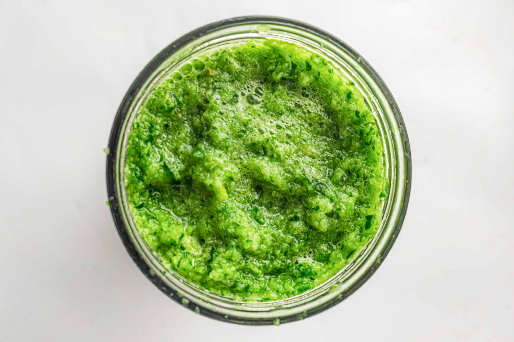 Overhead view of a jar filled with a green, chunky sauce or paste, likely made from blended herbs or vegetables, on a white background.