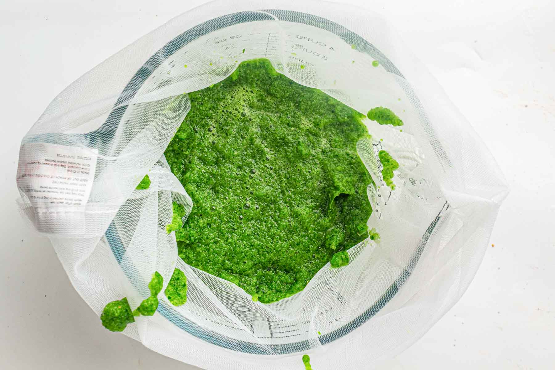 Bright green blended substance in a mesh strainer over a glass bowl, viewed from above, on a white surface.