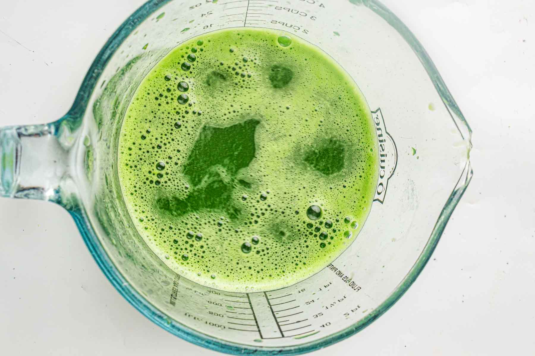 A glass measuring cup containing a frothy green liquid, viewed from above, with measurement markings visible on the side.