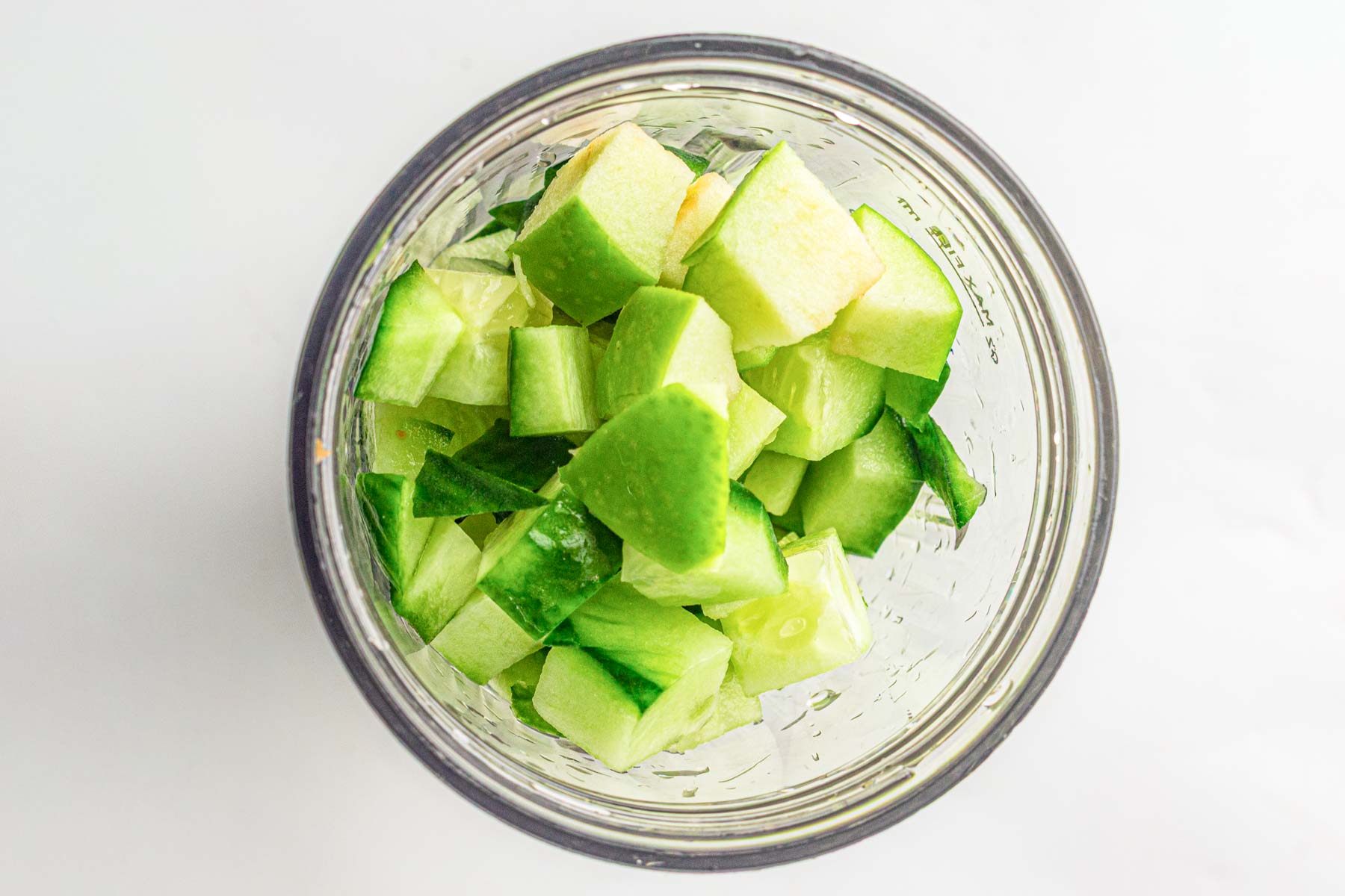 Chopped green apple and cucumber pieces in a glass blender jar, viewed from above on a white background.