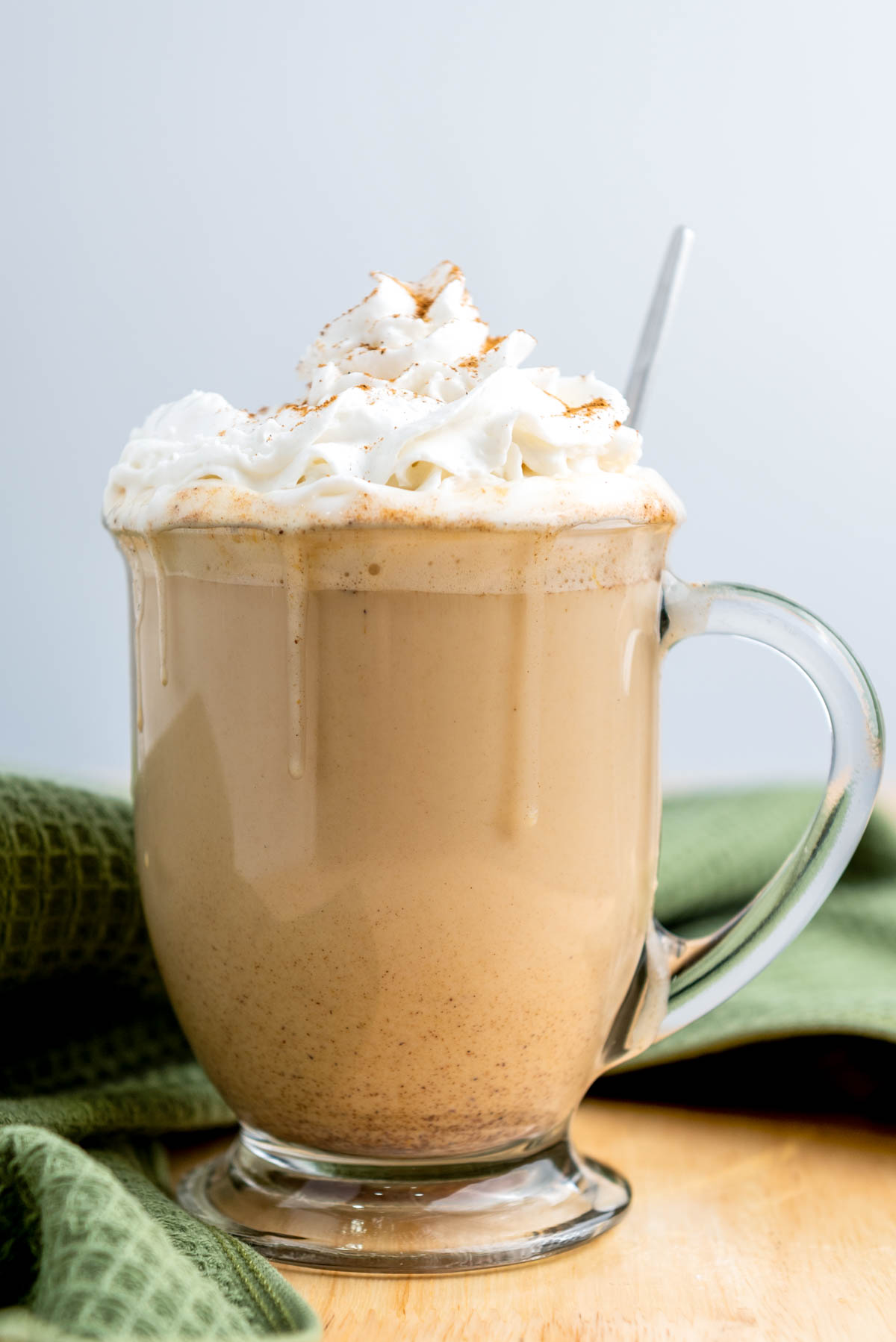 A glass mug filled with creamy coffee topped with whipped cream and a sprinkle of cinnamon, with a metal spoon, sits on a wooden surface next to a green cloth.